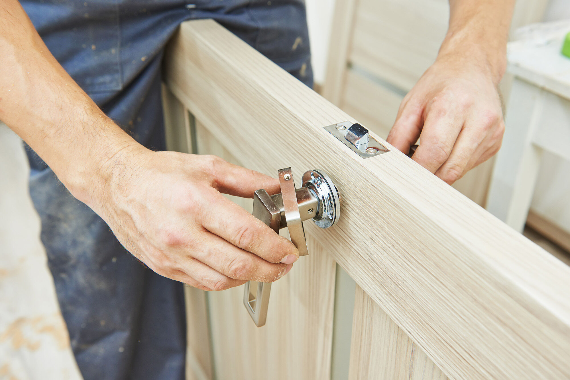 Nahaufnahme von Händen eines Handwerkers, der an einer hellen Holztür einen Türgriff mit Rosette montiert und ausrichtet. Oben im Türblatt ist das Schließblech beziehungsweise die Falle sichtbar, die Tür liegt schräg im Bild und wirkt noch unbehandelt oder neu. Im Hintergrund erkennt man eine Baustellenumgebung, das Motiv zeigt eine Tischler Kleinreparatur und die Montage von Türbeschlägen.