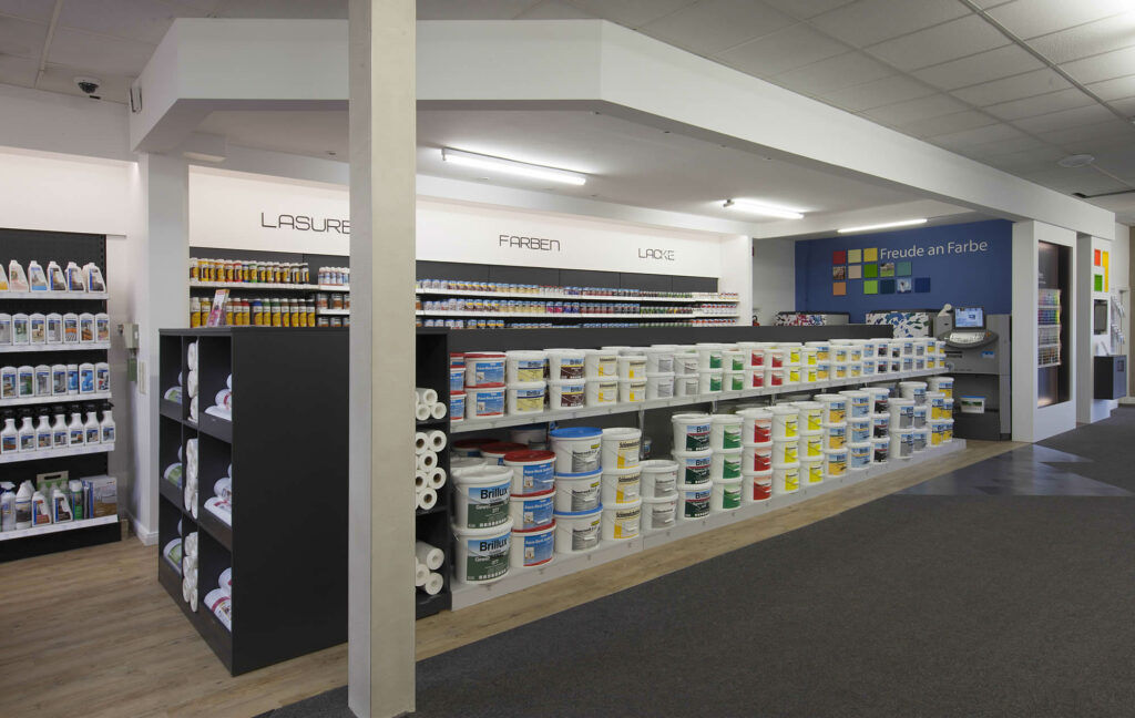 Heller Verkaufsbereich im Fachmarkt bei Ewering mit langen Regalwänden für Lasuren, Farben und Lacke. Vorne stehen viele große Brillux Farbeimer in mehreren Reihen auf niedrigen Regalen, dahinter sind an der Wand dicht sortierte Farbdosen und Gebinde auf langen Regalböden zu sehen.