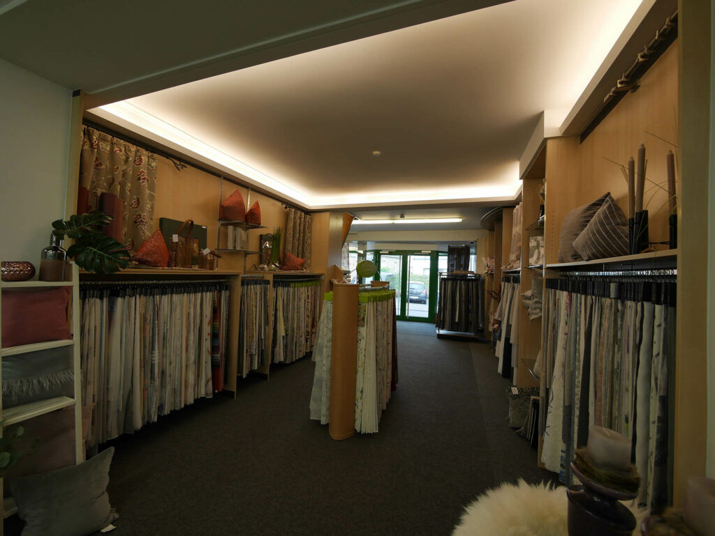 Gang in der Stoff- und Gardinenabteilung im Fachmarkt bei Ewering mit langen Reihen an Stoffmustern, die wie Gardinenmuster an Stangen hängen. Links und rechts stehen Holzregale mit vielen Musterhängern, darüber liegen Kissen, Deko und Stoffrollen, außerdem sind seitlich Vorhänge als Beispiel dekoriert. In der Mitte stehen weitere Musterständer, am Ende des Ganges sieht man die Eingangstür mit Blick nach draußen