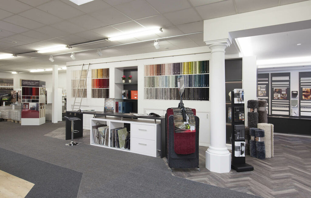 Großer Ausstellungsbereich im Fachmarkt bei Ewering mit einer weißen Musterwand, in der viele Teppichbodenmuster farblich sortiert in rechteckigen Feldern stecken. Davor steht ein langer schwarzer Beratungstisch mit einem Barhocker und einem weißen Unterschrank mit Musterbüchern. Links lehnt eine Leiter an der Musterwand, rechts stehen ein Musterständer mit Teppichmustern sowie mehrere aufgestellte Teppichboden-Proben, im Hintergrund sind weitere Ausstellungswände und Produktpräsentationen zu sehen