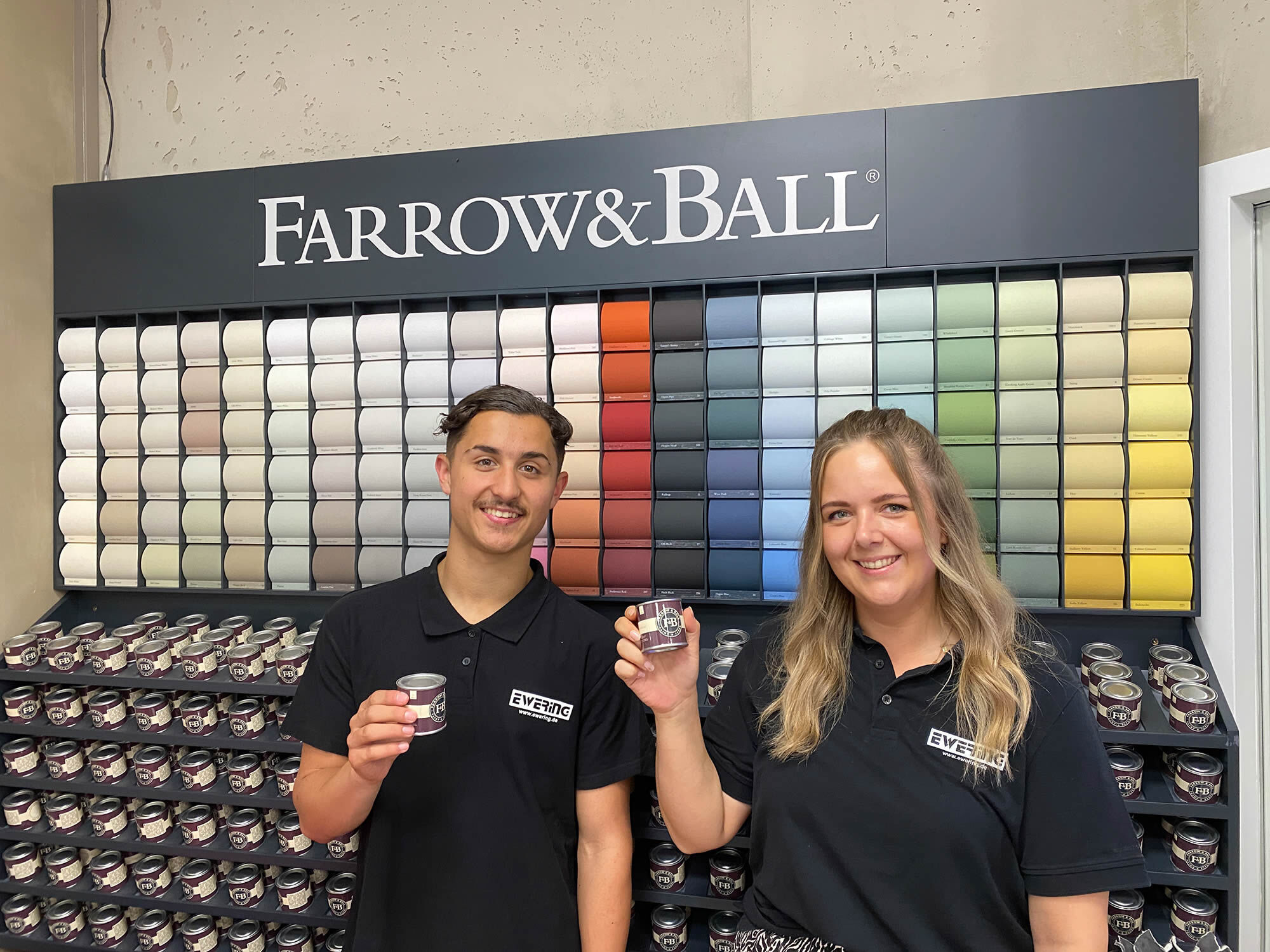 Zwei Ewering-Mitarbeitende im Fachmarkt vor der Farrow & Ball Farbwand mit Farbdosen und Farbkarten