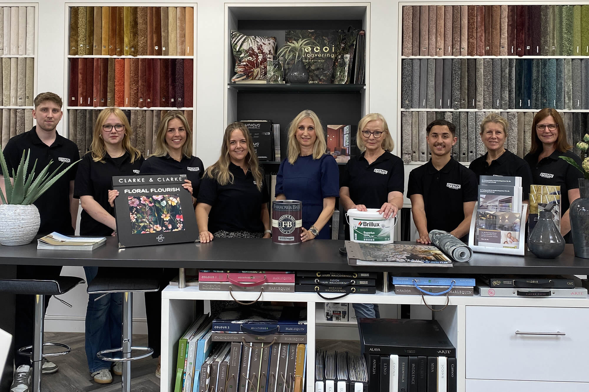 Gruppenfoto des Ewering Raumdesign Teams im Fachmarkt hinter einem großen schwarzen Beratungstisch. Mehrere Mitarbeitende stehen nebeneinander, alle in schwarzen Poloshirts mit Ewering-Logo, die Chefin trägt ein blaues Oberteil. Auf dem Tisch stehen ein großes Tapetenbuch „Clarke & Clarke Floral Flourish“, ein Farrow & Ball Farbeimer und ein Brillux Eimer, daneben liegen Tapeten- und Stoffmuster sowie ein gerolltes Musterstück. Im Hintergrund ist eine große Musterwand mit Teppichproben zu erkennen.
