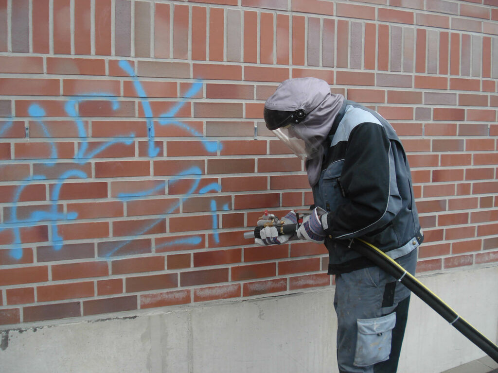 Mitarbeiter in Schutzkleidung entfernt blaues Graffiti von Klinkerwand - Untergrundreinigung