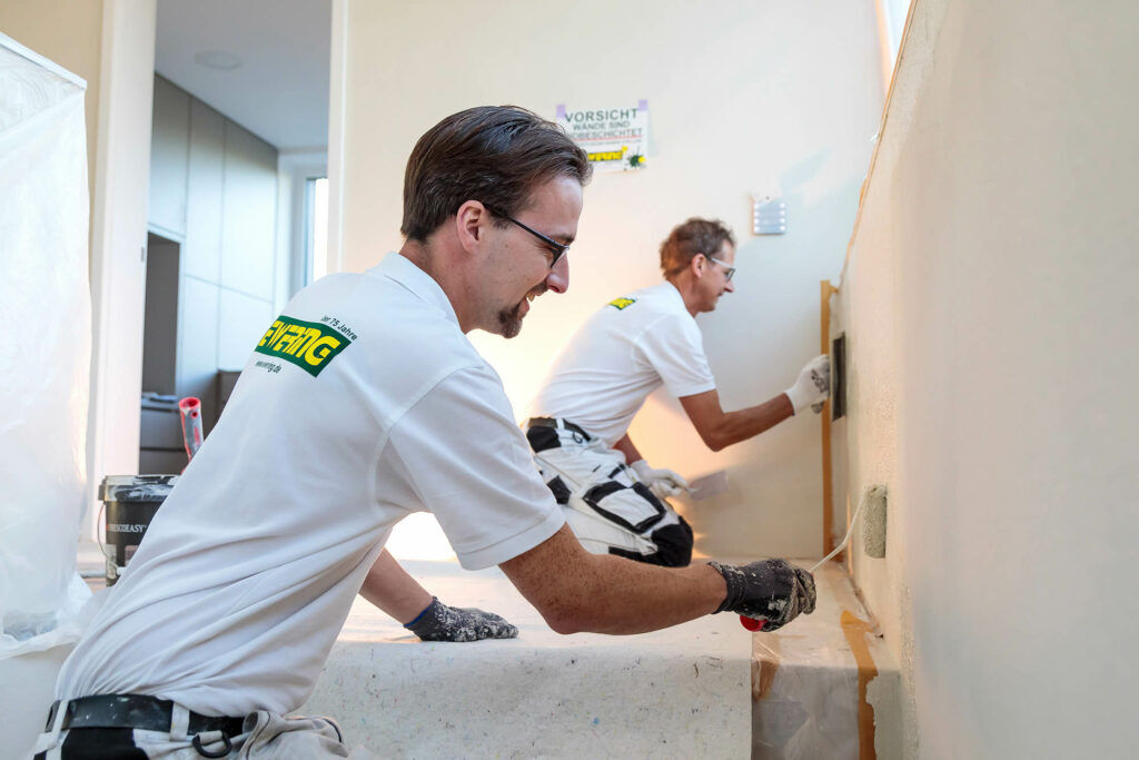 Zwei Ewering-Mitarbeiter im Polo-Shirt streichen konzentriert eine Wand auf Baustelle - Malerarbeiten