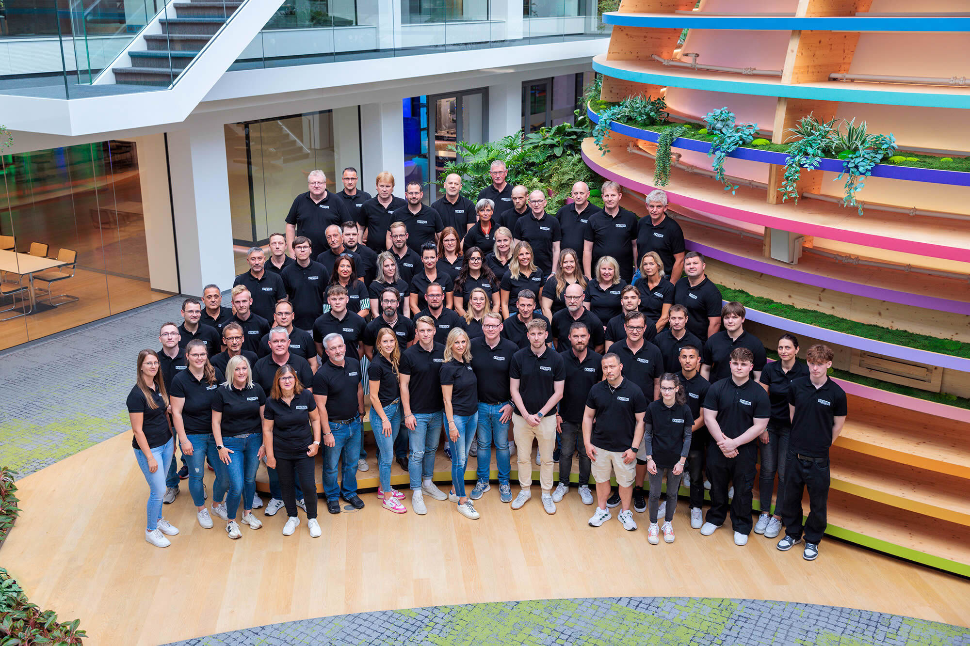 Großes Gruppenfoto des Ewering Teams in einem modernen, hellen Innenhof mit Glasfronten und offenen Fluren. Viele Mitarbeitende stehen in mehreren Reihen eng zusammen und tragen schwarze Poloshirts mit Ewering Logo, dazu überwiegend Jeans und Sneaker
