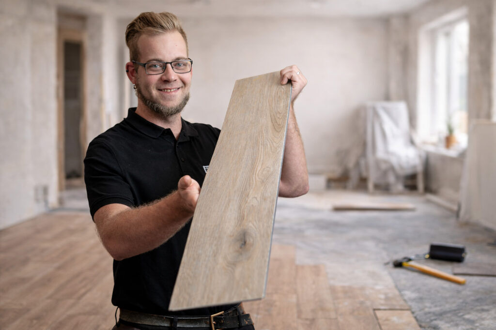 Junger Mann hält eine Bodenbelagsplanke in Holzoptik in einem renovierungsbedürftigen Innenraum