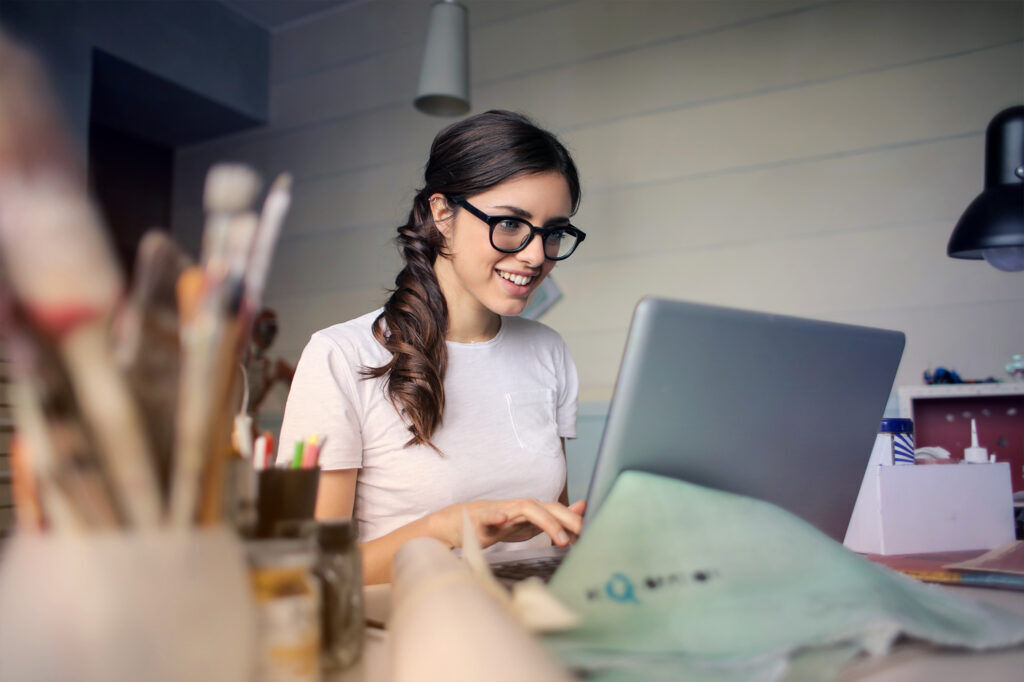 Junge Frau mit Brille arbeitet an einem Laptop an einem kreativen Arbeitsplatz mit Pinseln und Arbeitsmaterialien
