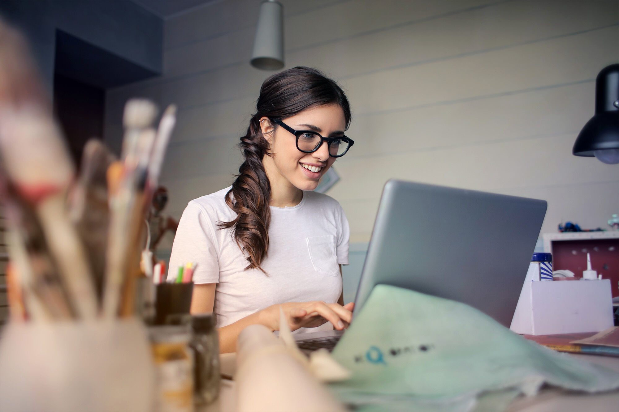 Junge Frau mit Brille arbeitet an einem Laptop an einem kreativen Arbeitsplatz mit Pinseln und Arbeitsmaterialien