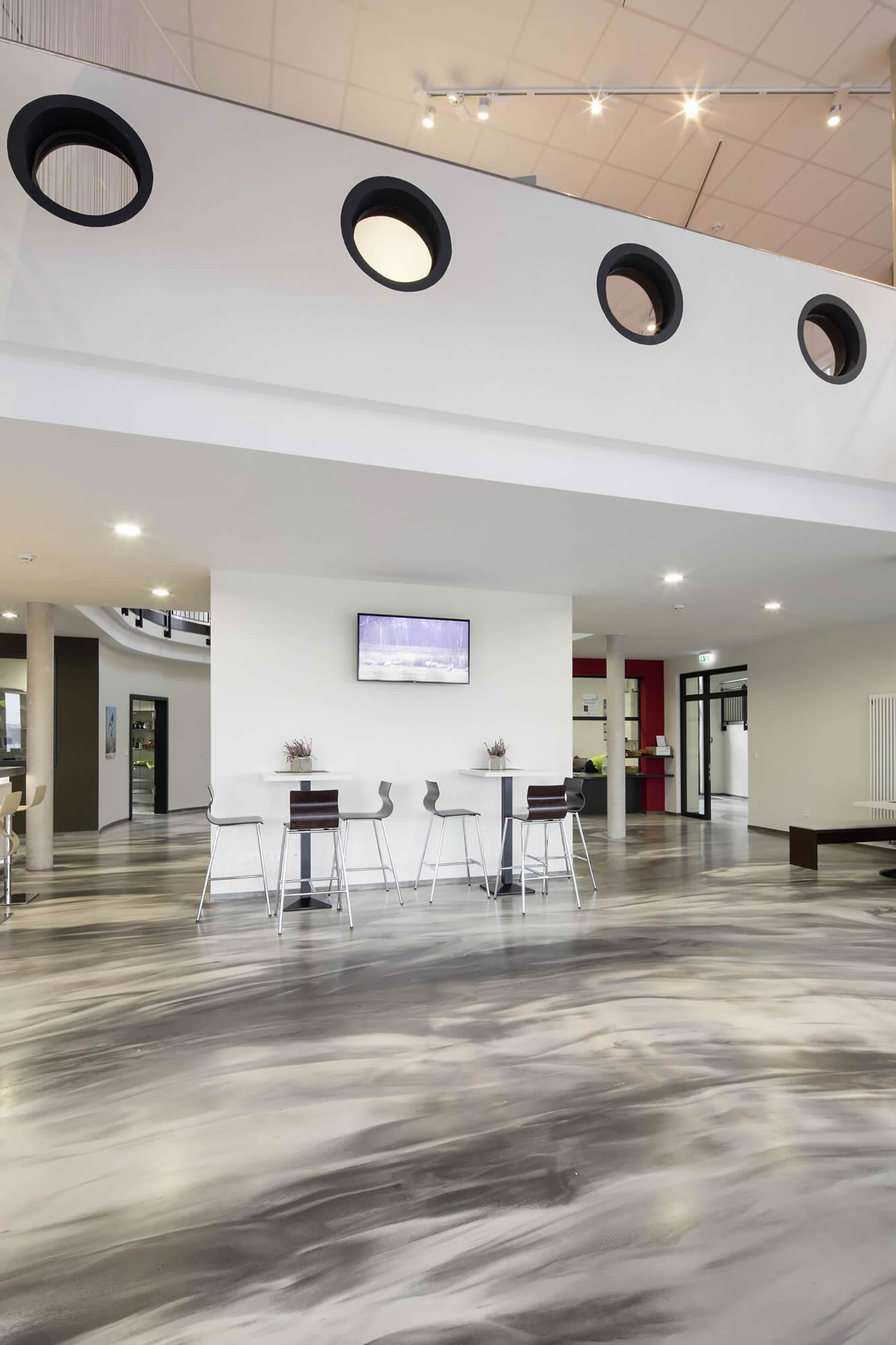 Großzügiger Innenraum mit Brillux Designboden in marmorierter Optik, hohen Tischen, Stühlen, Galerieebene und moderner Beleuchtung.