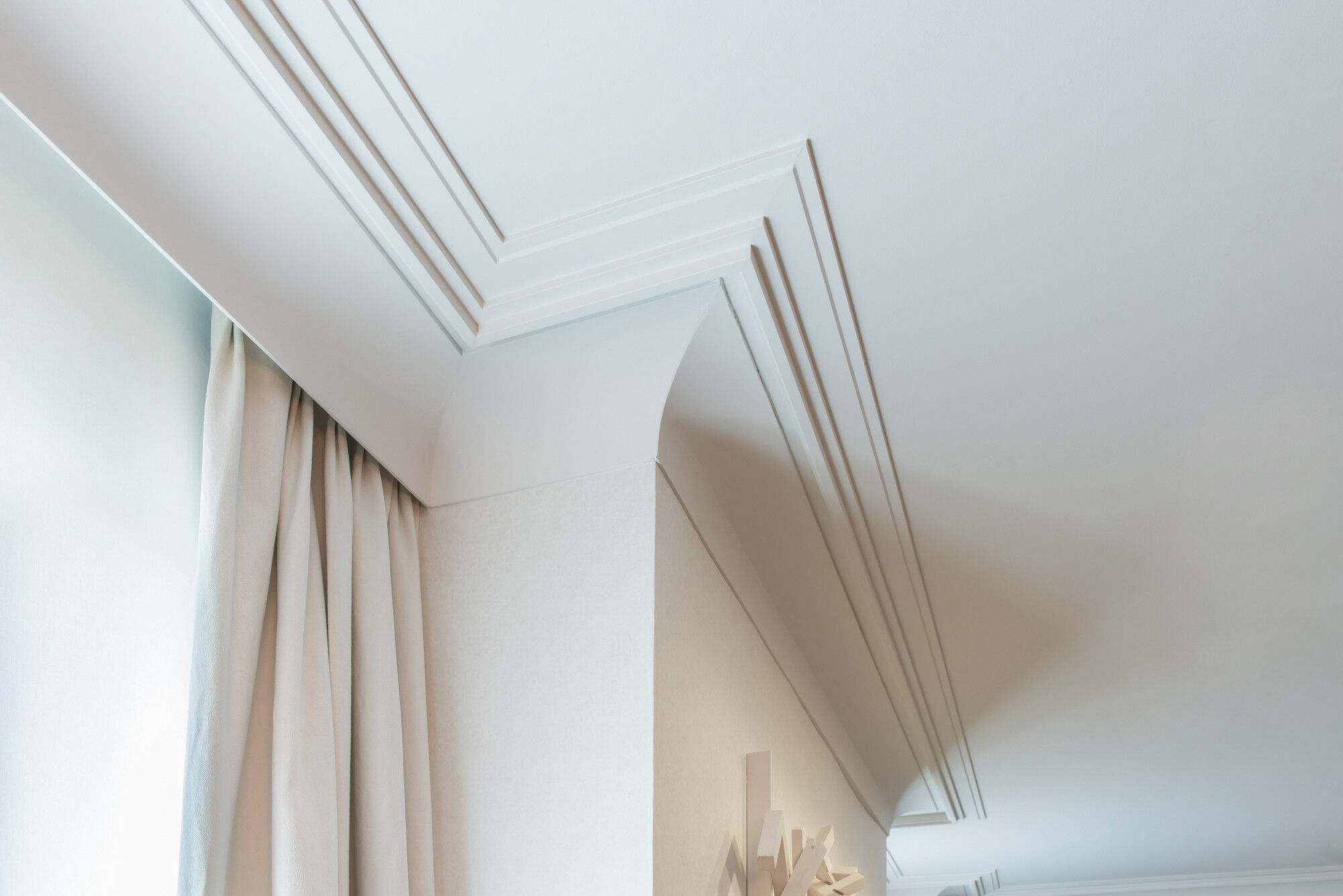 Detailaufnahme einer weißen Deckenleiste und Stuckleiste mit klarer Profilierung an einer hellen Decke neben einem Vorhang