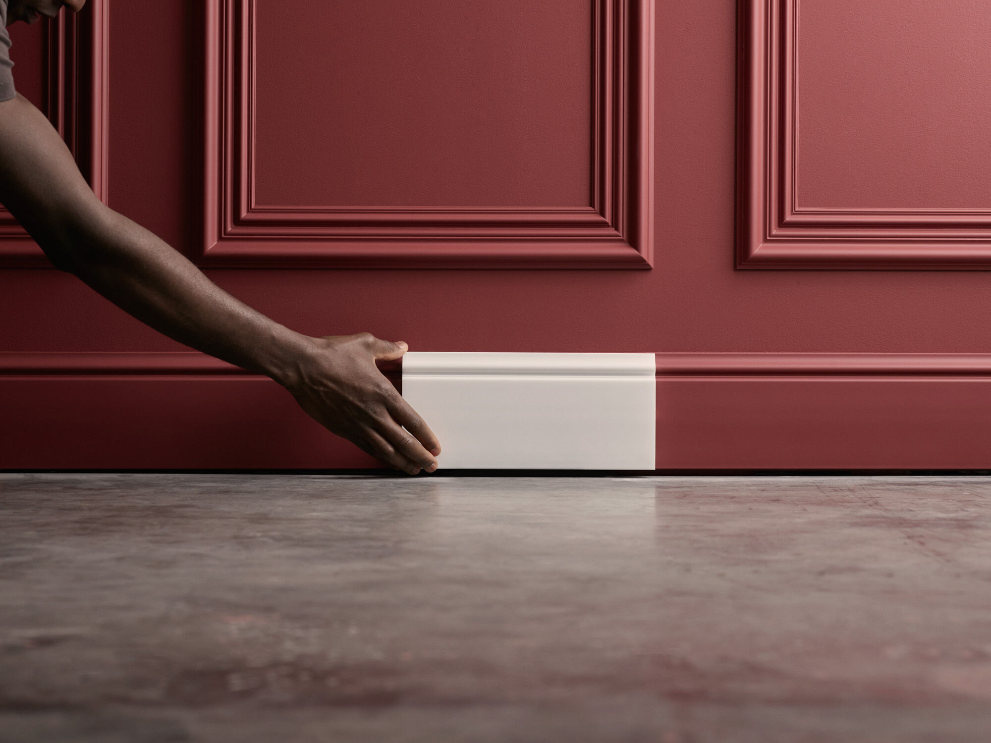 Hand setzt eine weiße Fußleiste an eine dunkelrote Wand mit profilierten Wandfeldern und klassischer Wandgestaltung