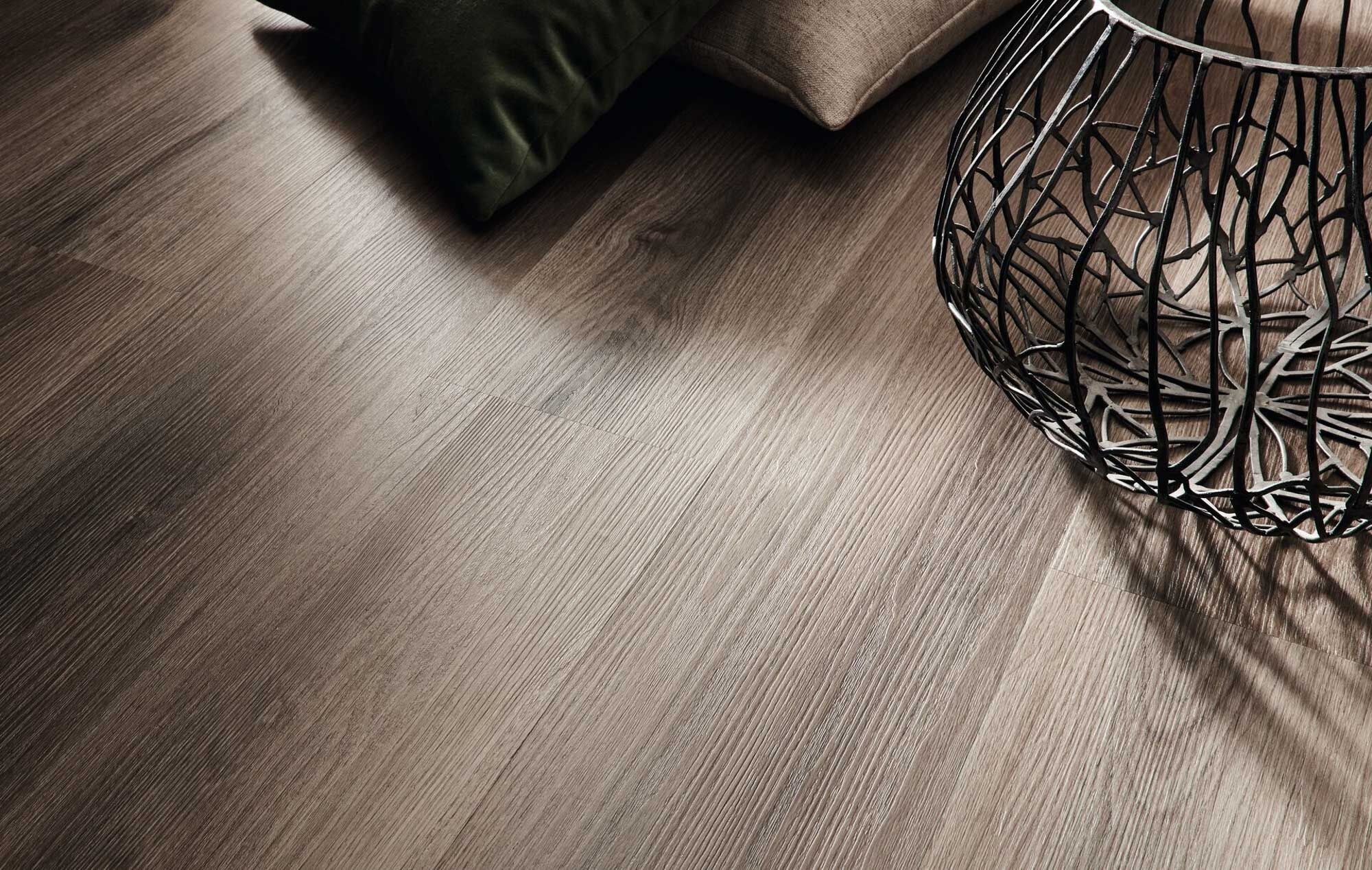 Detailaufnahme eines dunkelbraunen Designbelags in Holzoptik mit Dielenformat, feiner Maserung und schwarzem Metallbeistelltisch