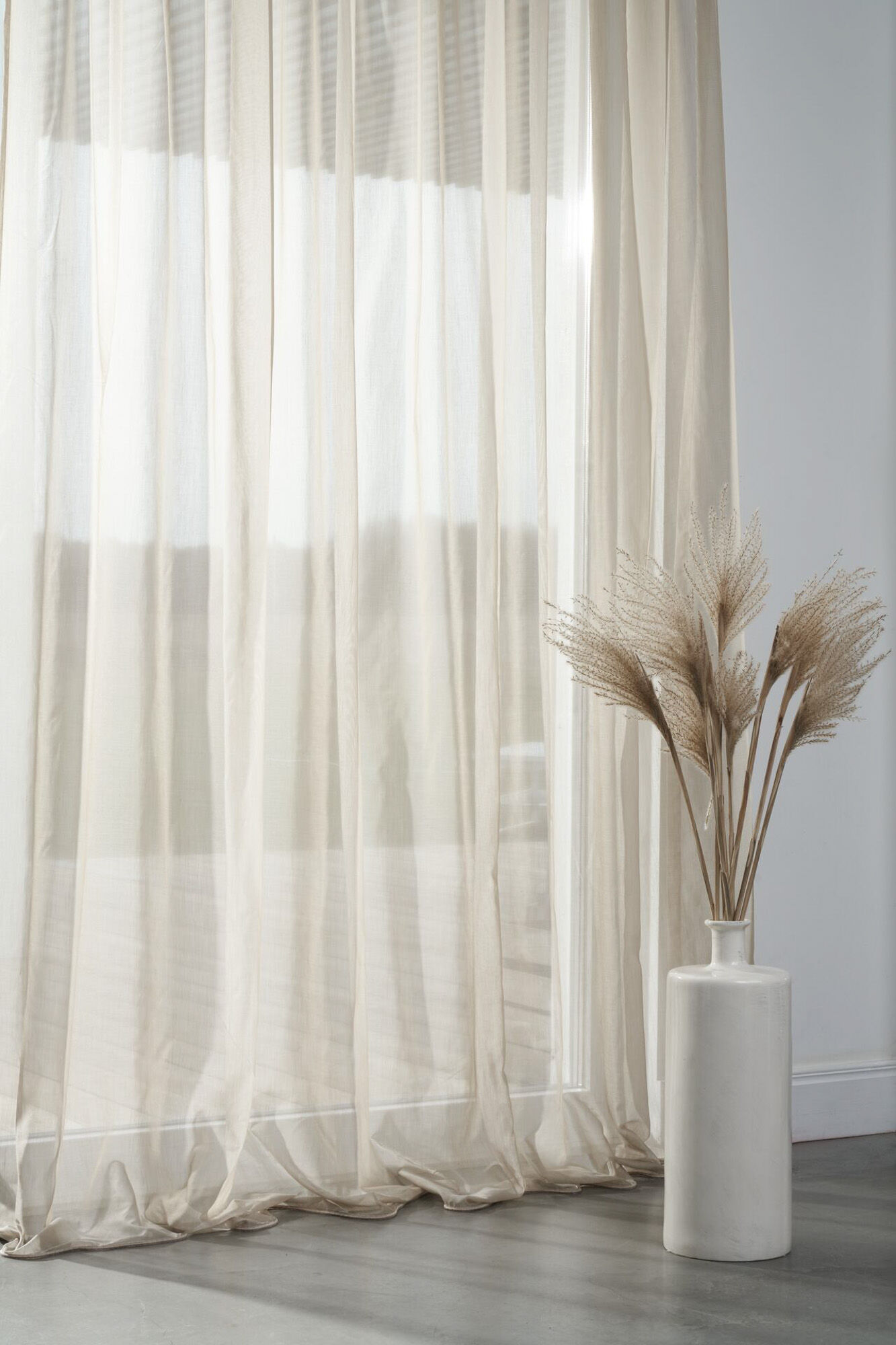 Bodenlange, transparente Gardine in hellem Naturton vor einer Fensterfront mit weichem Faltenfall und dekorativer Vase