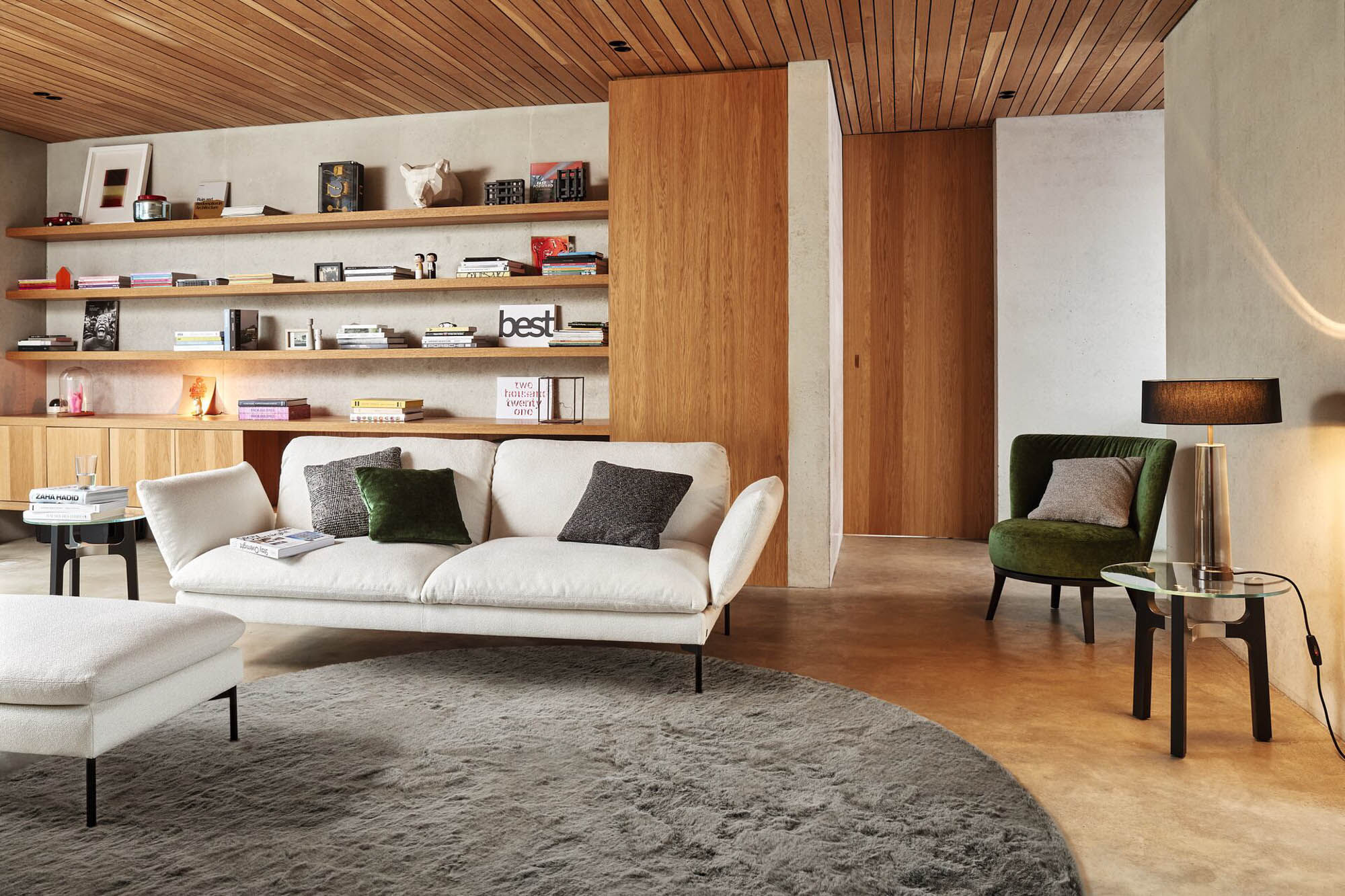 Modernes Wohnzimmer mit rundem grauem Hochflor-Teppich, hellem Sofa, grünem Polstersessel, offenen Holzregalen und Holzdecke