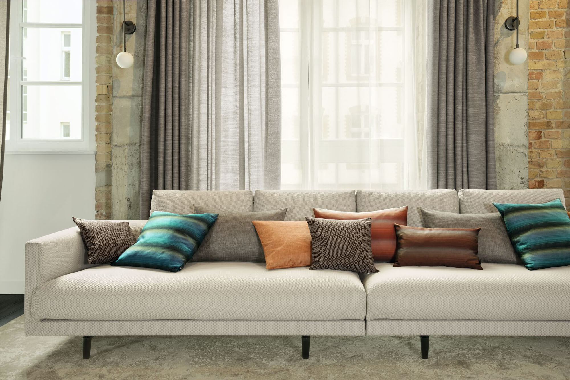 Modernes Sofa mit hellem Polsterbezug, dekorativen Kissen, bodenlangen Vorhängen, transparentem Store und dezentem Teppich von JAB ANSTOETZ