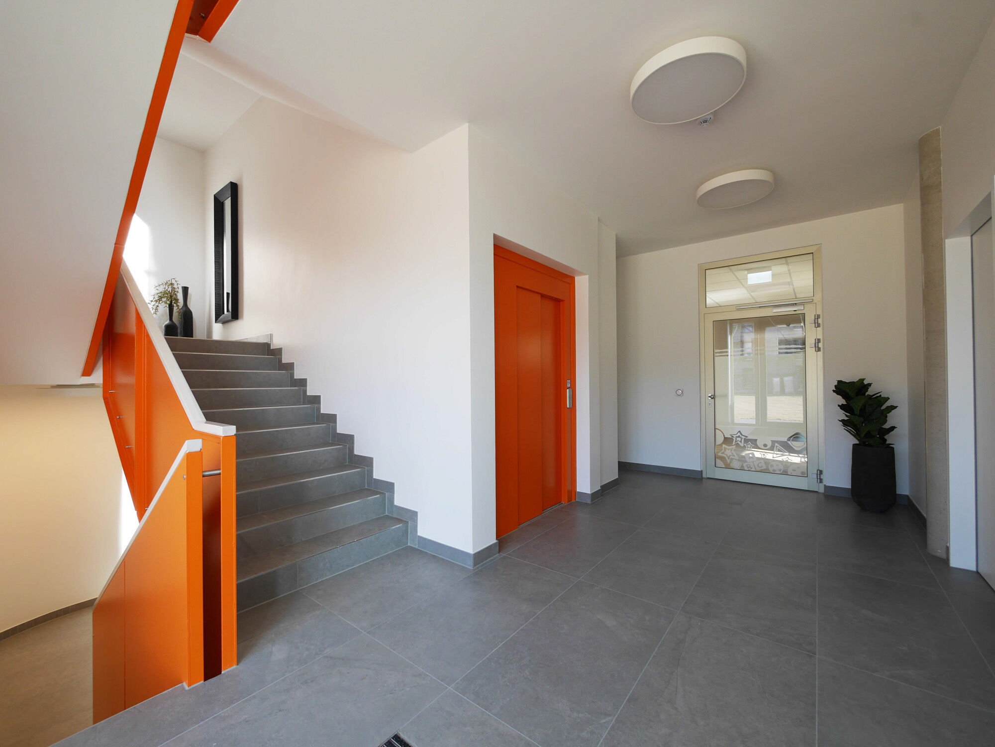 Treppenhaus mit orange-roten Treppenkanten, dunkelgrauen Stufen und Eingangsbereich mit Pflanzen – IDPM Gebäude B