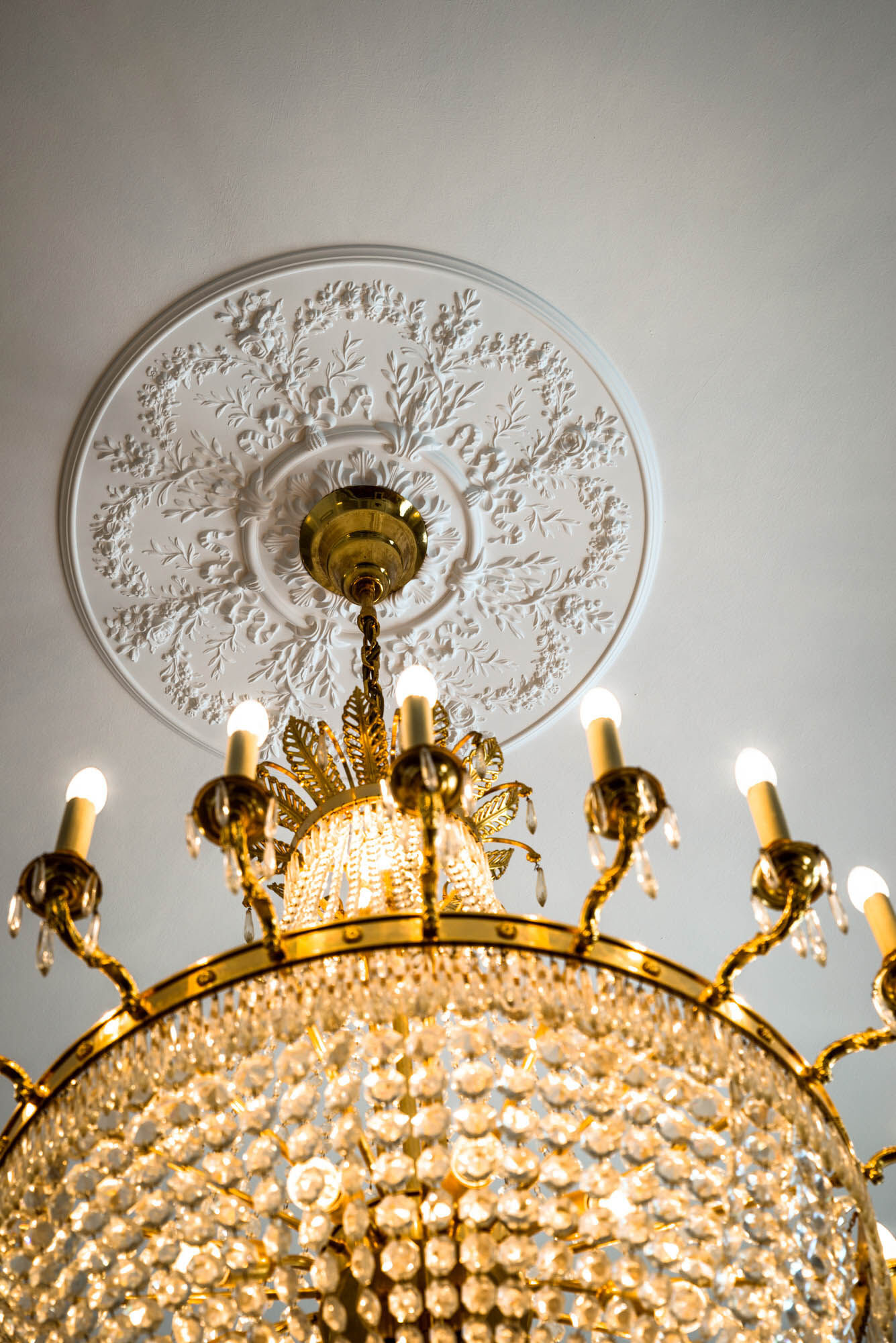 Weiße Deckenrosette mit floraler Ornamentik und goldfarbenem Kronleuchter an einer hellen Decke