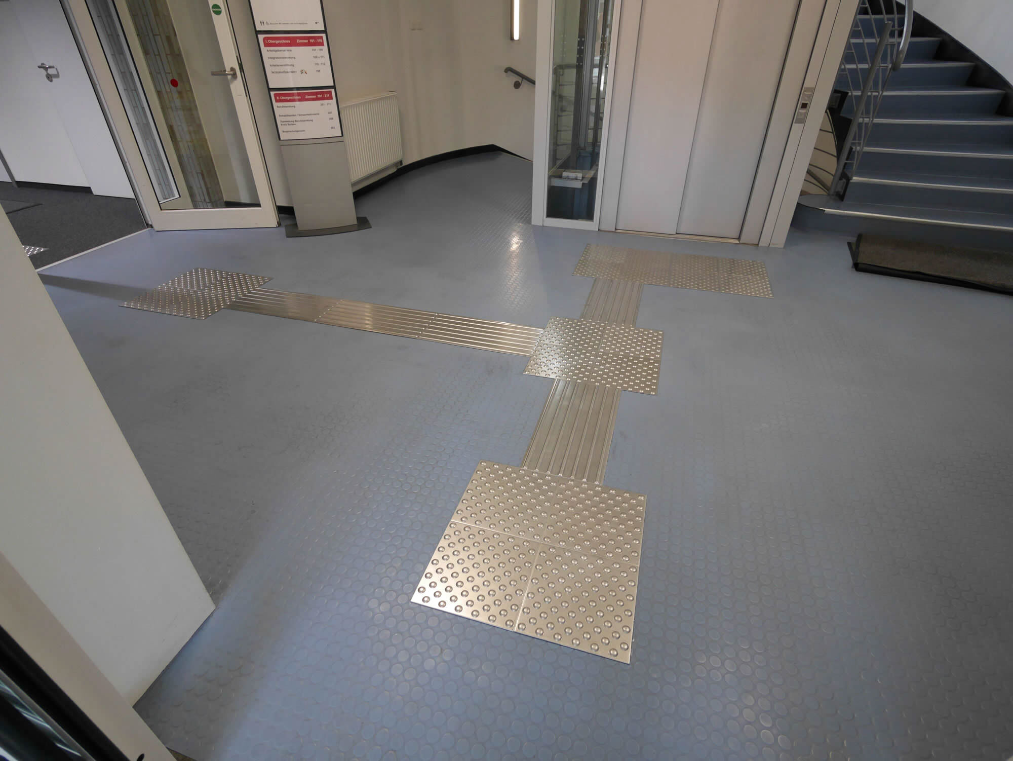 Blindenleitsystem aus Edelstahl mit Leitstreifen und Noppenfeldern vor Aufzug und Treppe.