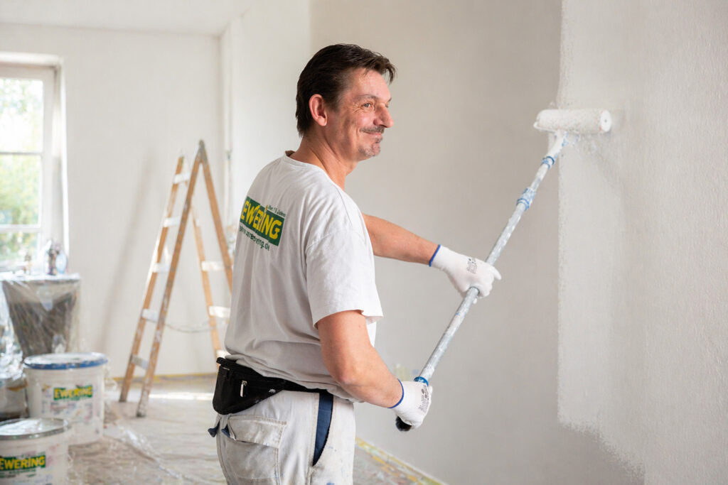 Ewering Mitarbeiter streicht eine weiße Innenwand mit Farbrolle in einem vorbereiteten Raum