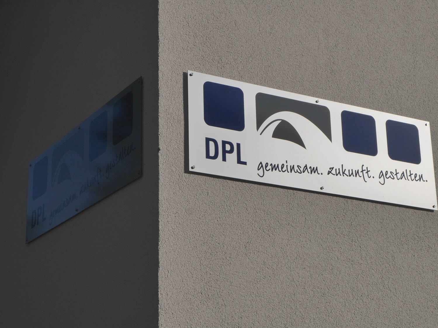 Helles DPL-Fassadenschild mit Brückenmotiv an grauer Hauswand - Grafikdesign Werbetechnik Fassadenschild