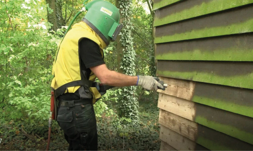 Mitarbeiter mit grünem Strahlhelm reinigt verwitterte Holzfassade - Untergrundreinigung