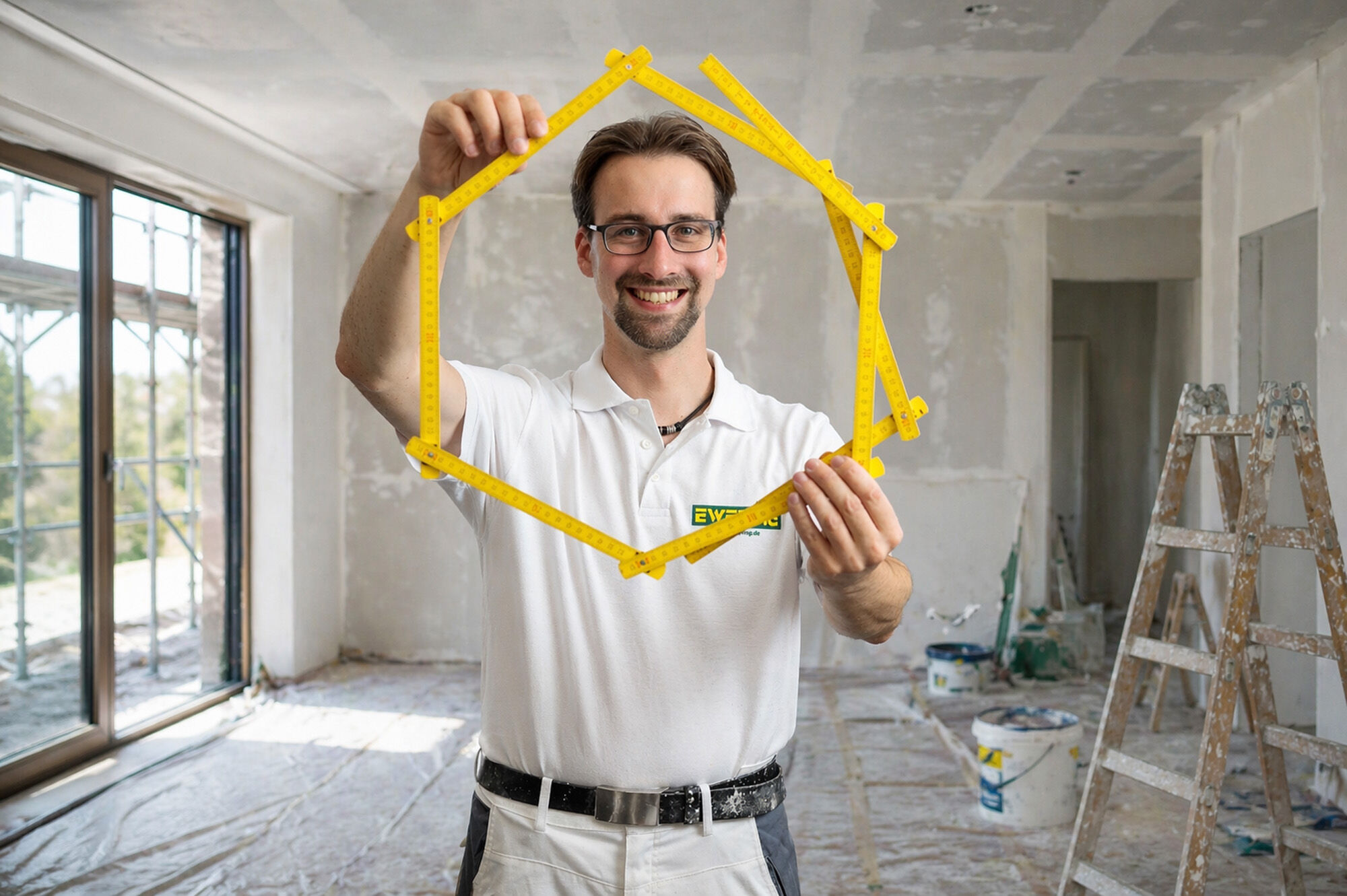 Maler und Lackierer von Ewering hält einen Zollstock in Hausform auf einer Baustelle