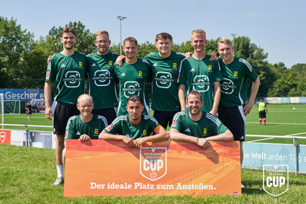 Das Team Ewering nimmt am Firmenfußballturnier Volksbank Gescher Cup in Gescher teil und steht gemeinsam in grünen Trikots auf dem Sportplatz vor dem Turnierschild